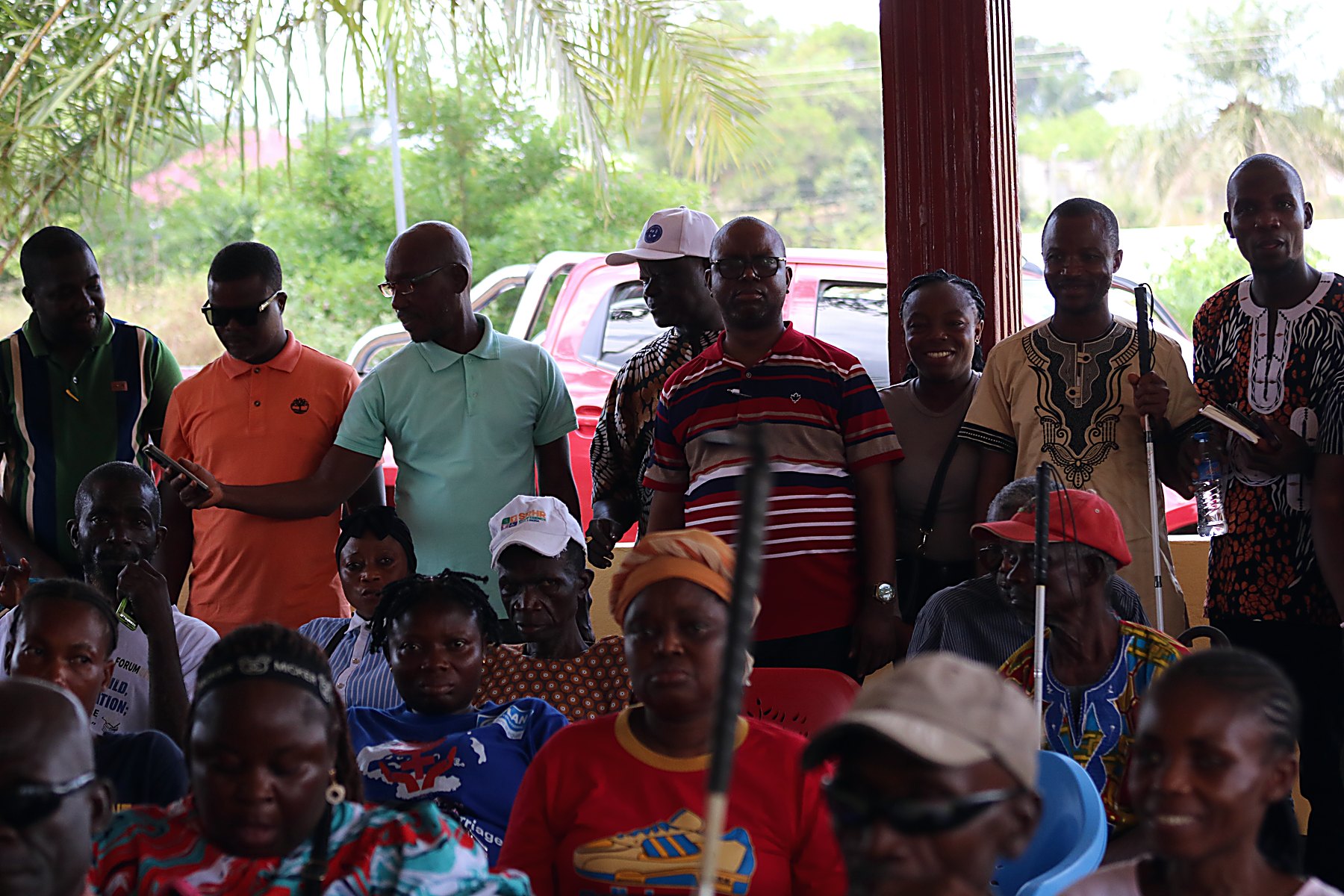 A crowd of persons with disabilities and community members gathered under a pavilion, several holding white canes and crutches.