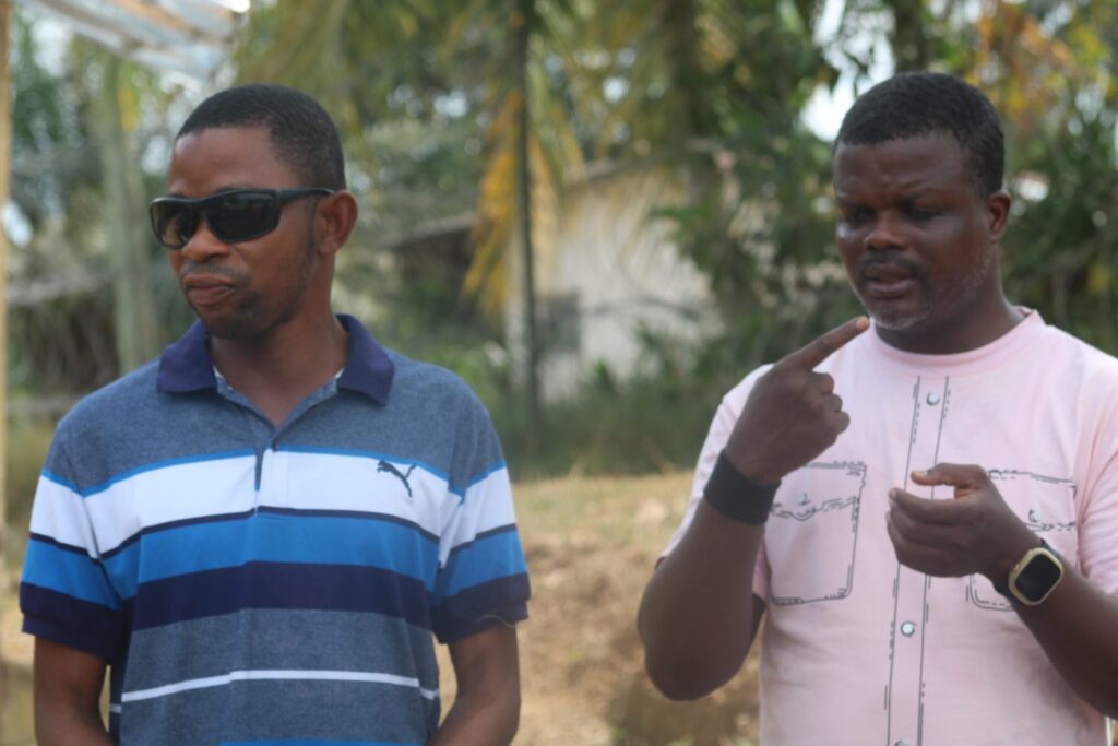Two men in conversation outdoors, one signing and one wearing dark glasses.