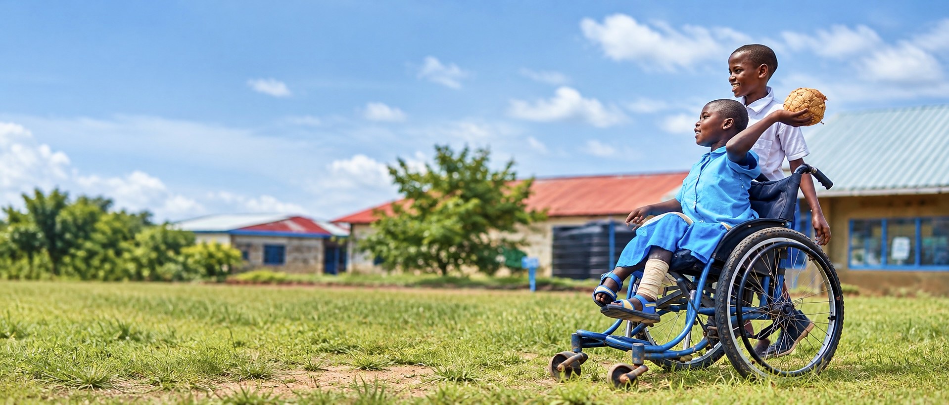 A boy pushes a classmate who uses a wheelchair across a school field, both of them smiling.