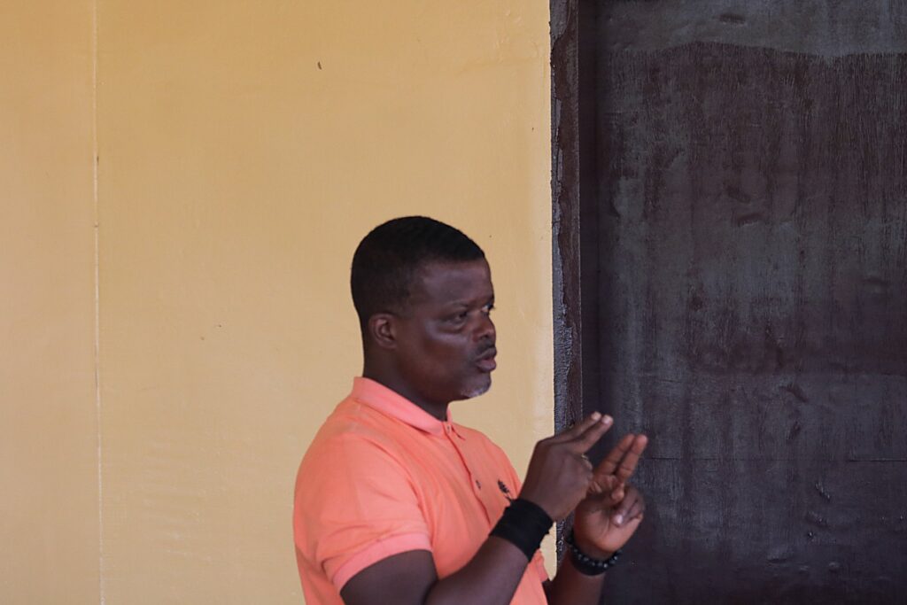 A man demonstrating a sign in Liberian sign language against a plain wall.
