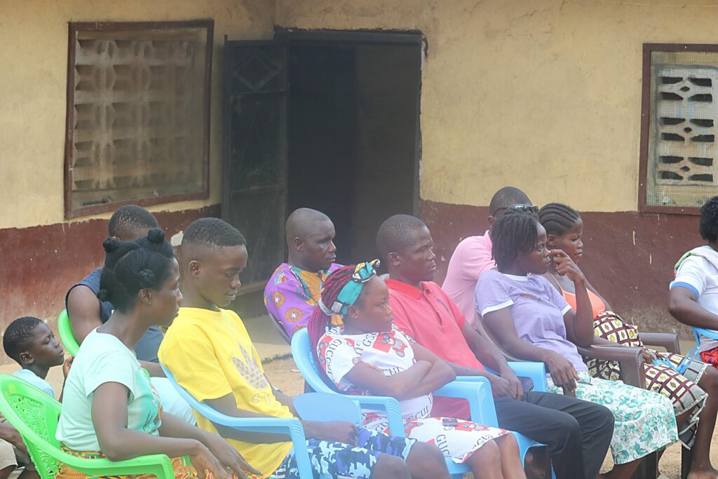 Community members seated on plastic chairs outside a building during a village consultation.