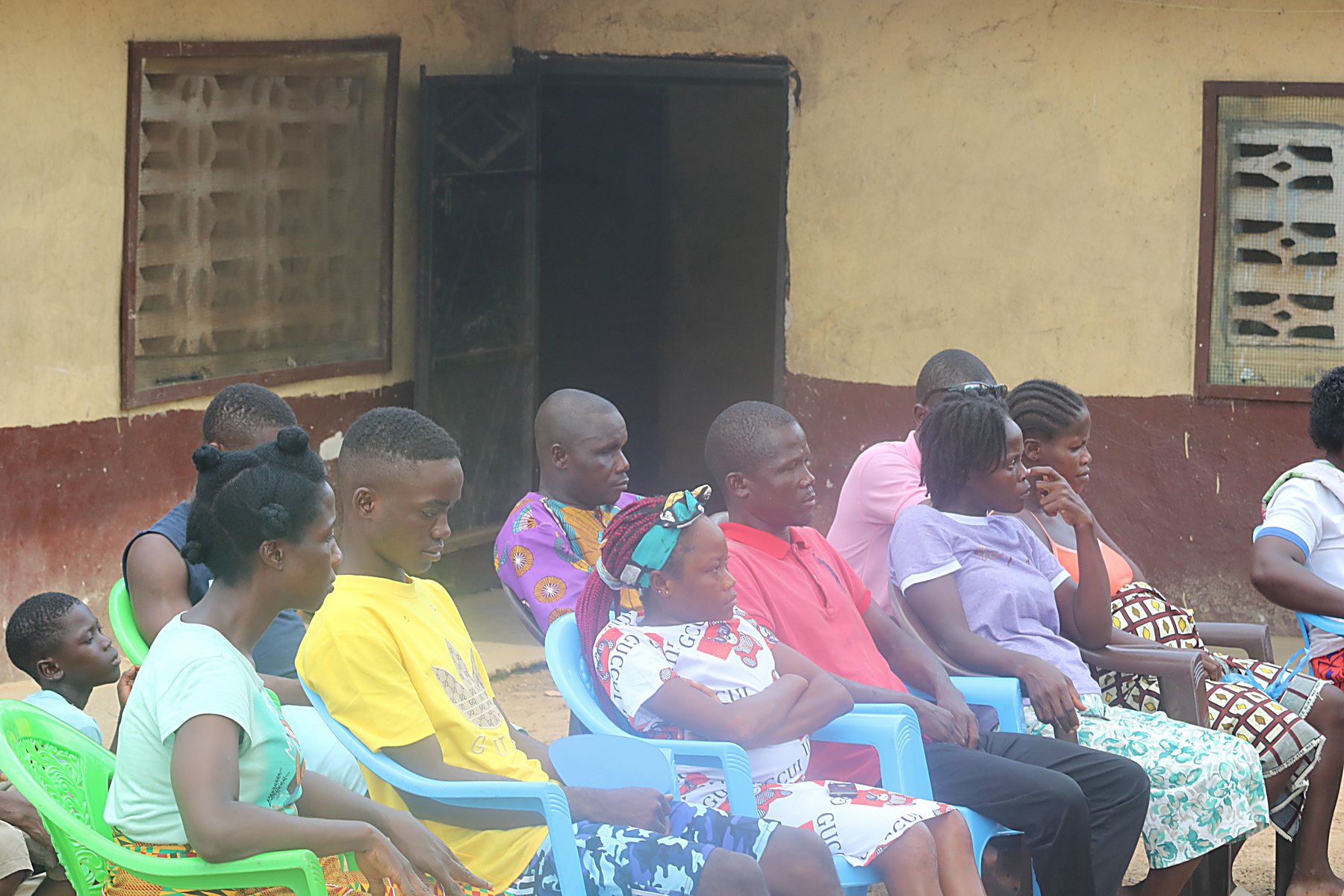 Community members seated on plastic chairs outside a building during a village consultation.