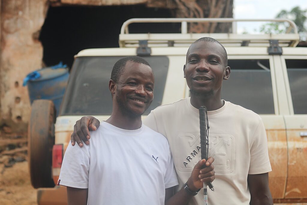 Two men standing together beside a Commission vehicle, one holding a white cane.